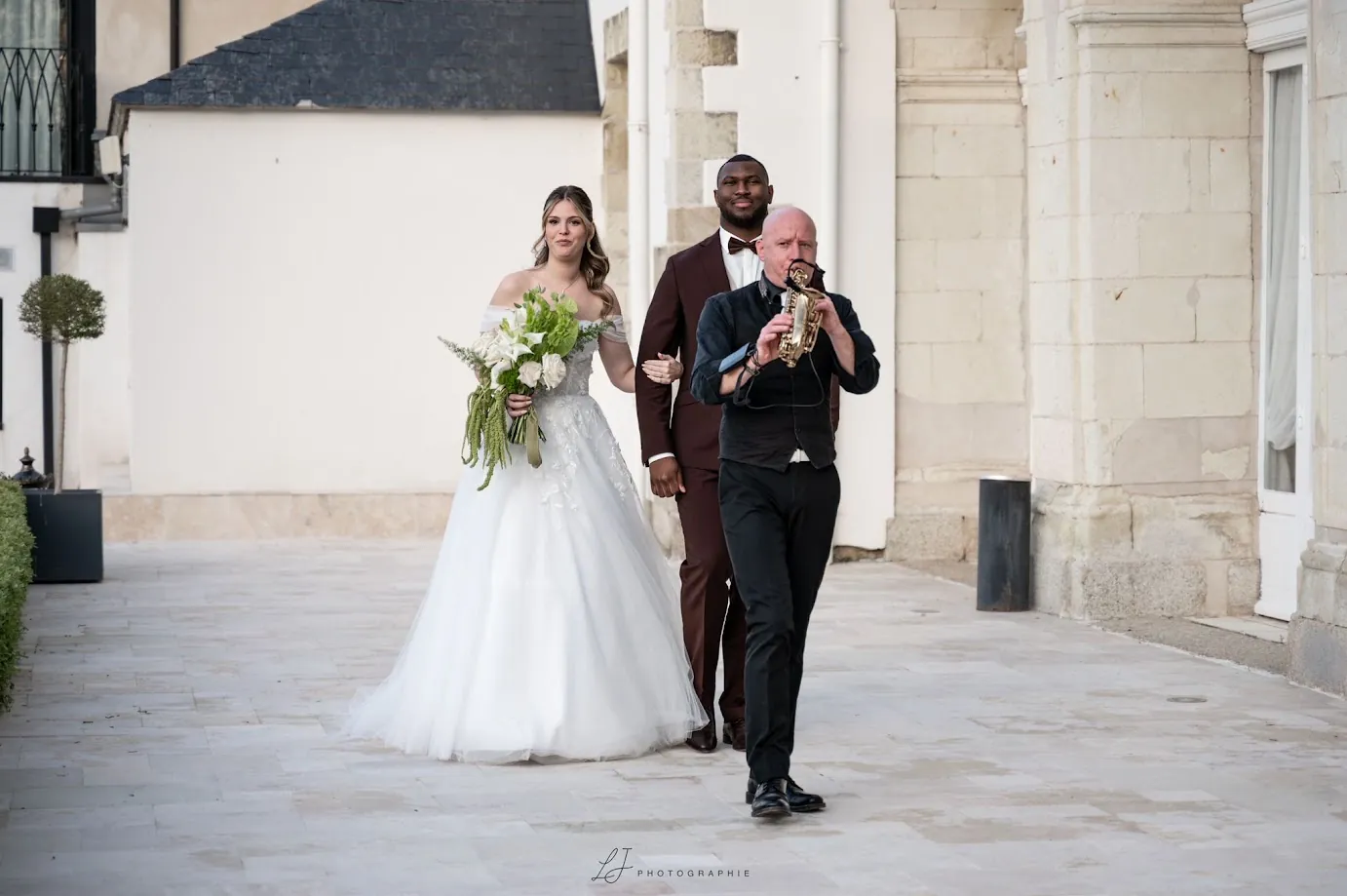 Marine, un bouquet d’arums et de feuillage dans les mains, et Yacouba, en costume bordeaux, avancent sur une terrasse de pierre derrière un saxophoniste