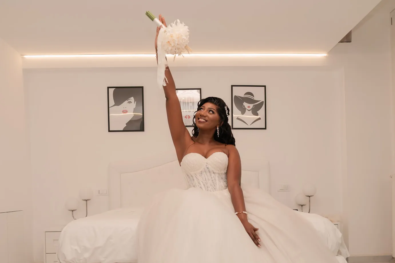 Dans une chambre blanche, Naomie en robe de tulle, assise sur le lit, lance son bouquet de fleurs blanches vers le ciel