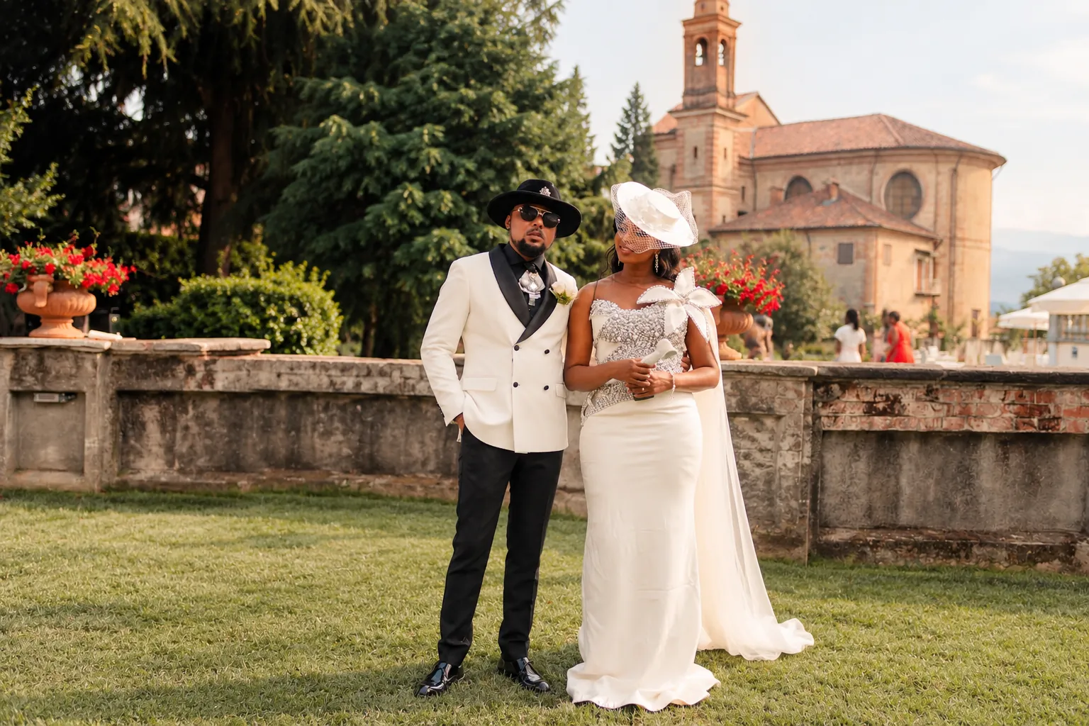 Naomie et Patrick posent côte à côte devant un muret de pierre, dans les jardins de la Villa Merlin : lui en veste blanche à revers noirs et chapeau, elle en robe blanche et voilette, avec l’église de brique en arrière-plan
