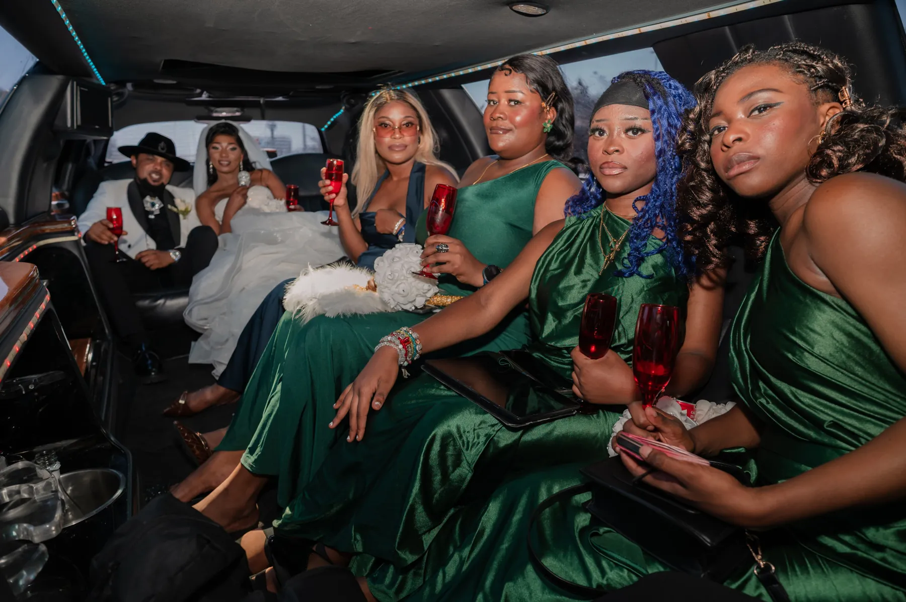 Dans une limousine, les mariés et leurs demoiselles d’honneur en robes vertes lèvent des coupes rouges