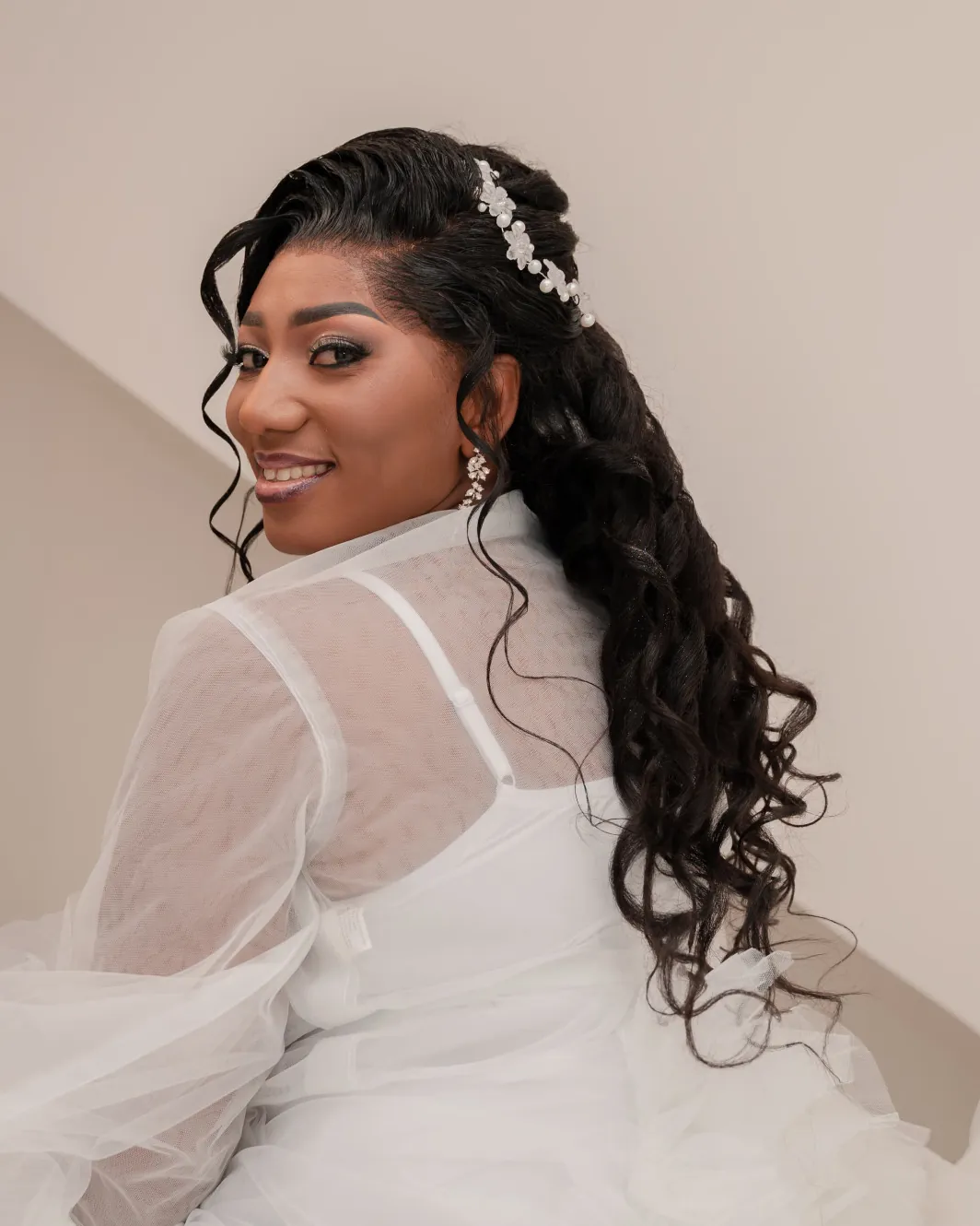 Naomie pendant ses préparatifs, en robe de tulle blanc, tournée vers l’objectif et souriante, une barrette de perles dans les cheveux