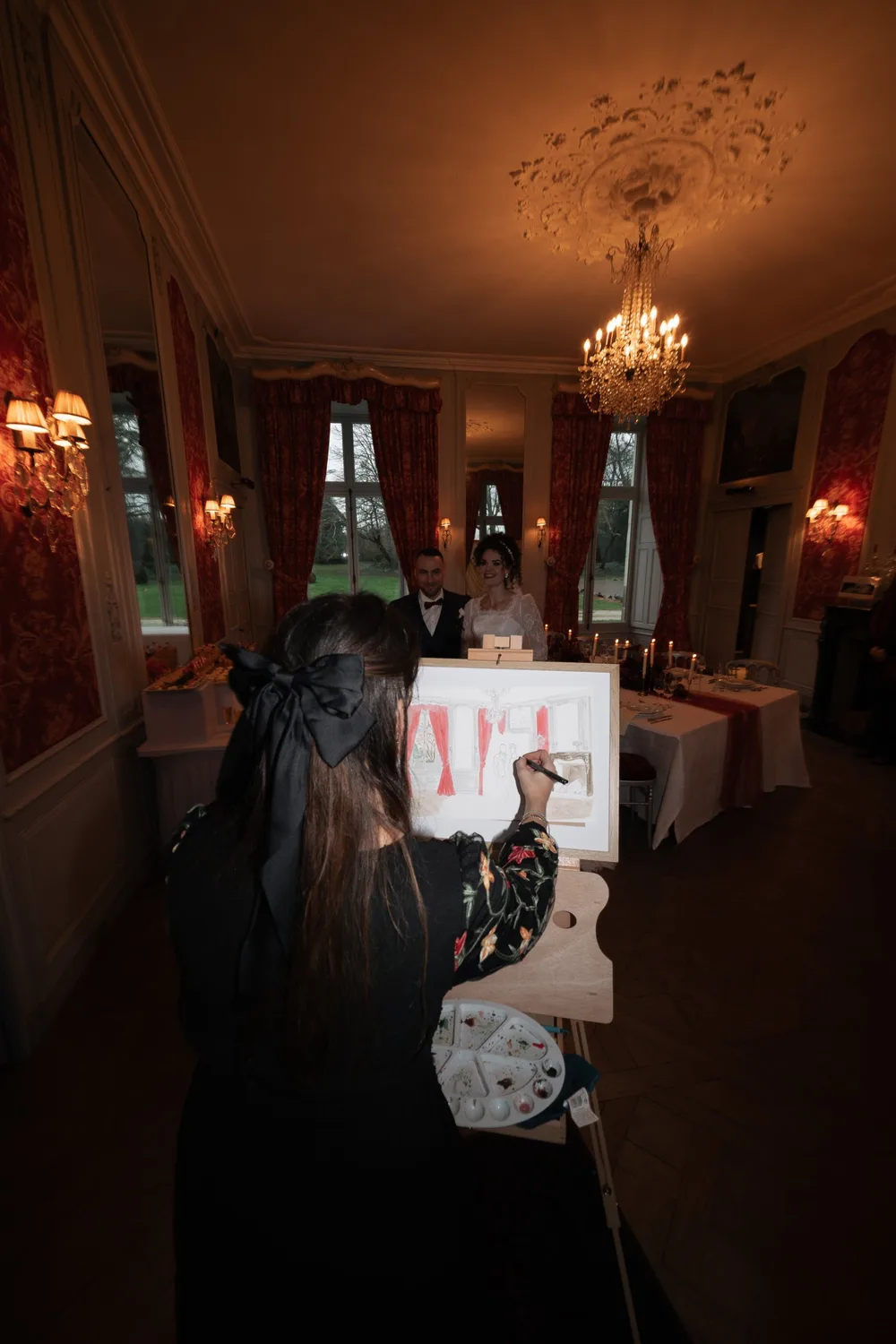 Dans un salon de château aux boiseries et aux rideaux rouges, une artiste peint la scène sur son chevalet, face aux mariés qui la regardent en souriant