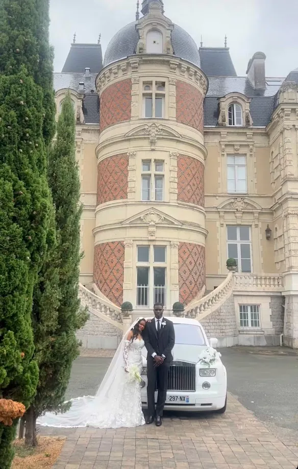 Au loin, les mariés devant la façade en brique et pierre du château, à côté d’une voiture blanche fleurie, entre un cyprès et la cour pavée