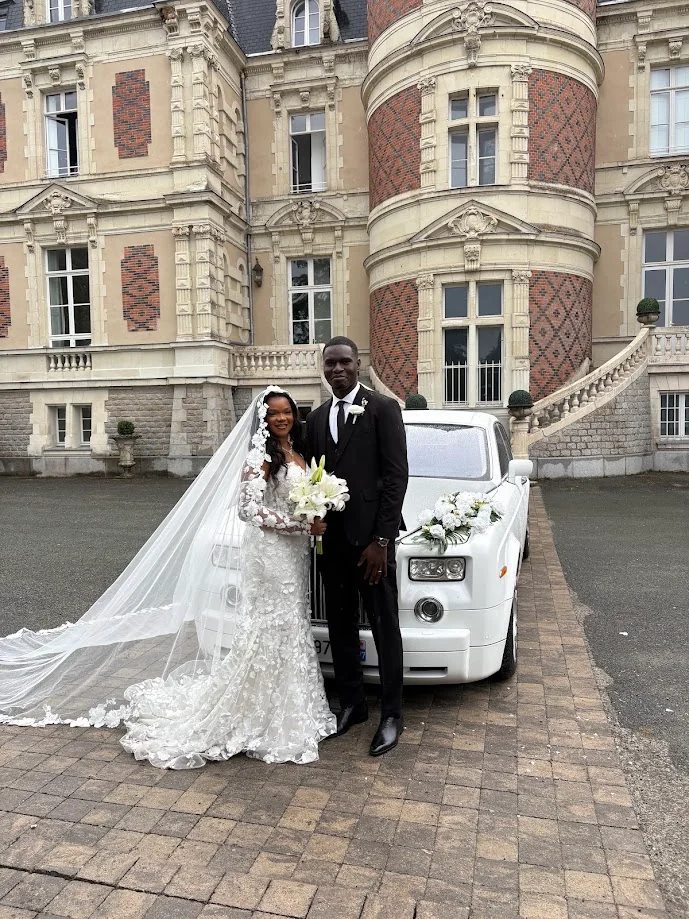 Pepita et David posent devant une voiture ancienne blanche ornée de fleurs, face à la façade en brique et pierre d’un château