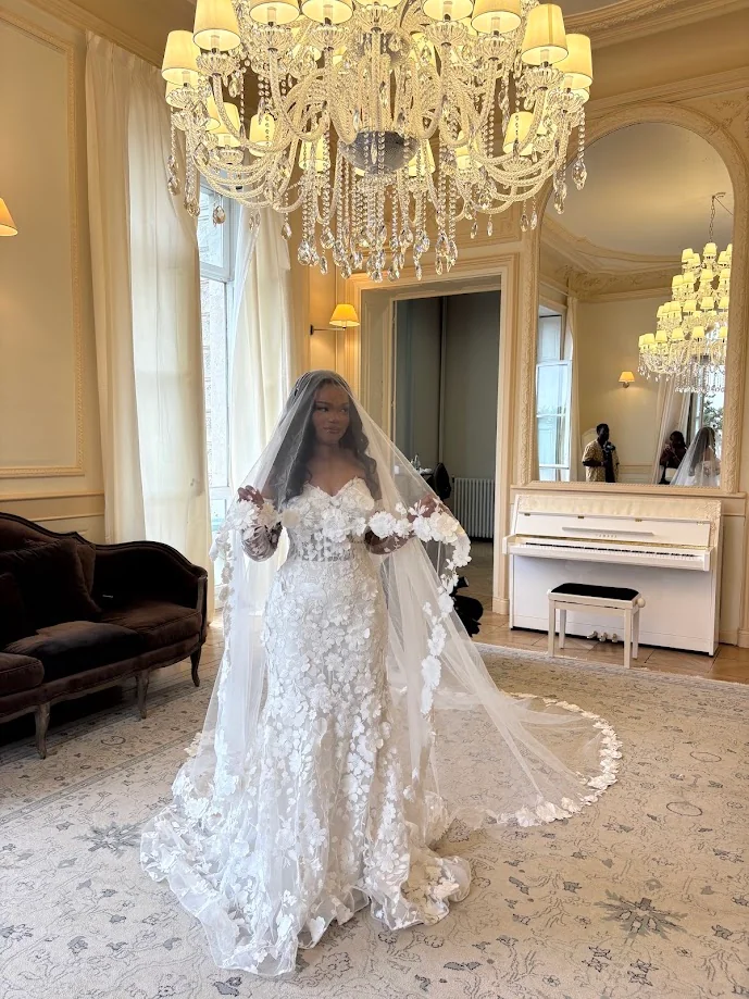 La mariée, en robe de dentelle fleurie et long voile, au centre d’un salon sous un lustre de cristal, près d’un piano blanc