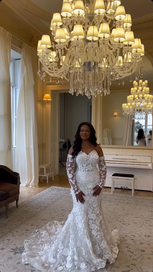 La mariée, en robe de dentelle fleurie, debout sur un tapis clair dans un salon sous un lustre de cristal, près d’un piano blanc