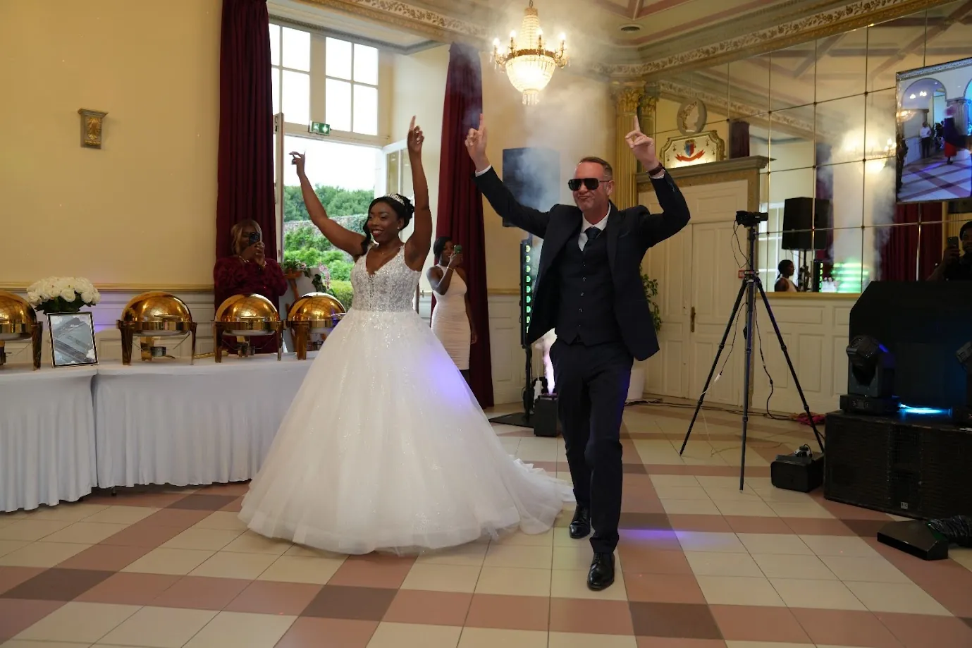 Dans la salle de réception, Rose, la robe étalée, et Jean-Luc, les bras levés et lunettes de soleil, ouvrent la piste de danse
