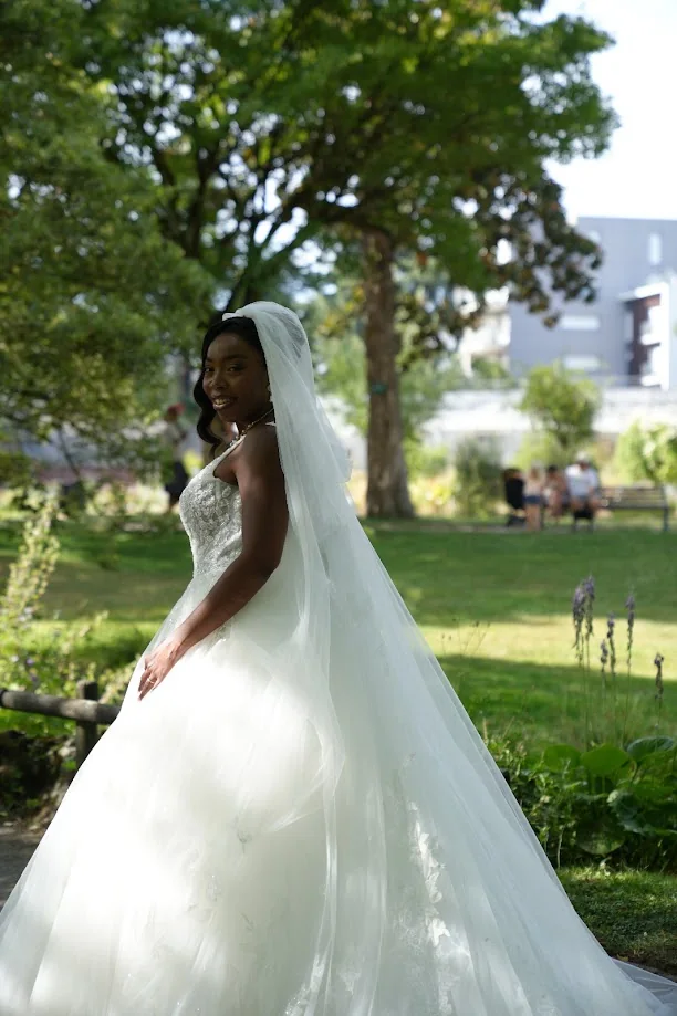 La mariée, de profil et souriante, sous un long voile de tulle, dans un parc