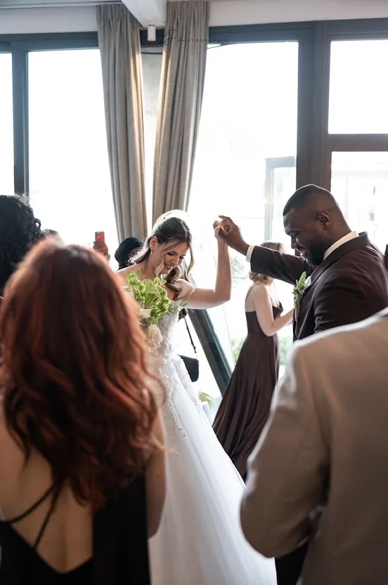 La mariée, son bouquet à la main, entre dans la salle en souriant ; le marié lui tend la main, les proches autour d’eux
