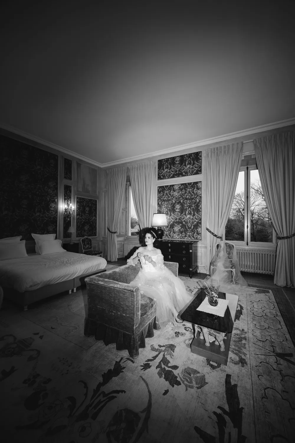 En noir et blanc, une chambre de château lumineuse : lit, fauteuil, grandes fenêtres drapées et tapis, la mariée assise au fond de la pièce
