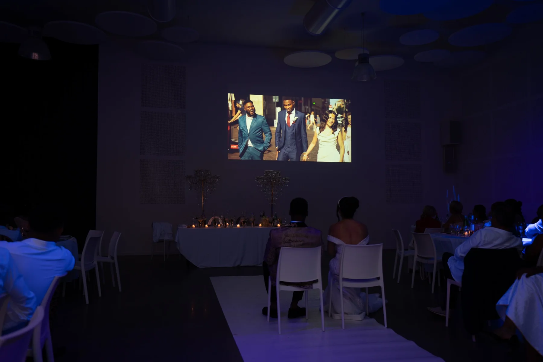 Pendant la réception, les mariés, vus de dos, regardent le film de leur histoire projeté sur grand écran dans une salle plongée dans une lumière bleutée