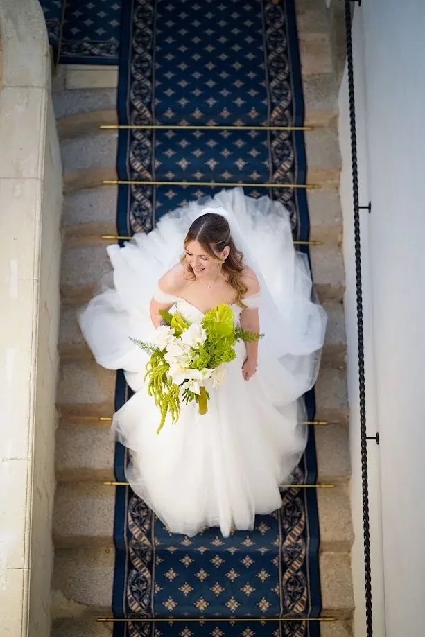 La mariée descend un escalier au tapis bleu, son bouquet vert et blanc à la main, vue de dessus