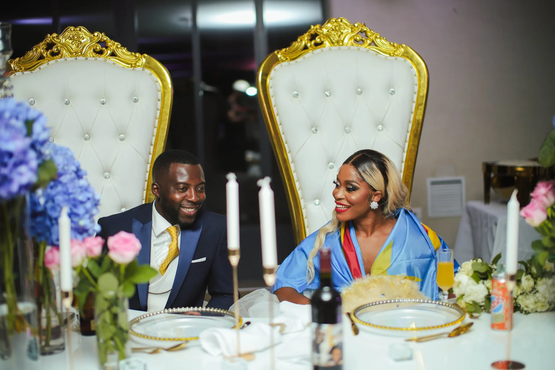 Les mariés, assis à leur table d’honneur sur deux grands fauteuils blancs et dorés, échangent un sourire complice parmi les bougies et les hortensias