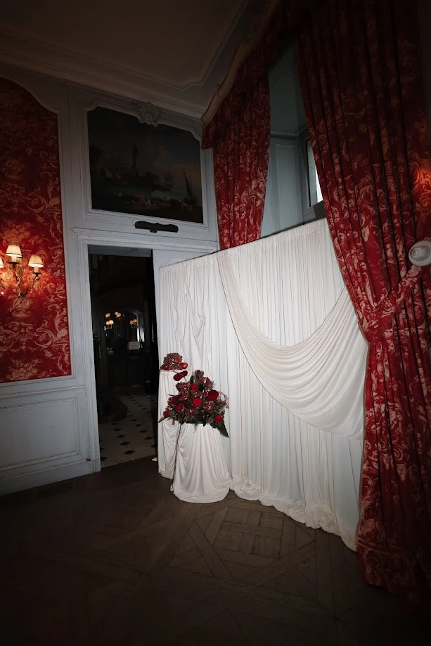 Dans un salon de château aux tentures de toile rouge, un grand drapé blanc et une composition de fleurs rouges posée sur un piédestal drapé